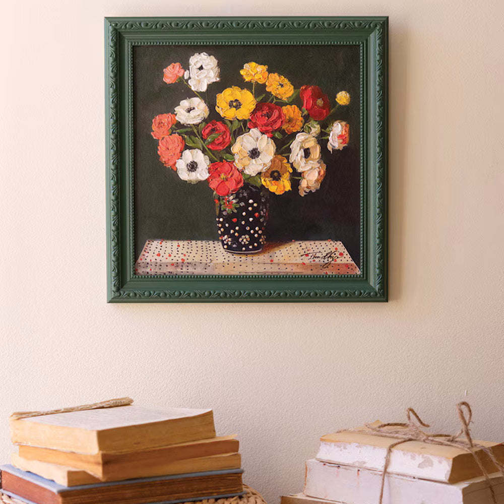 Still life oil painting of colorful flowers in a polka dot vase with a green frame, hanging above stacks of vintage books.