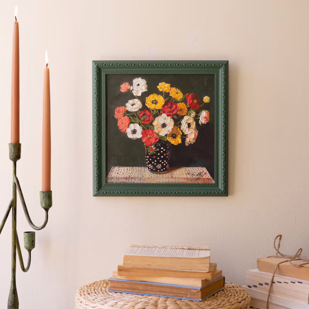 Still life oil painting of colorful anemones in a polka dot vase, featured in a green frame on a cream wall.