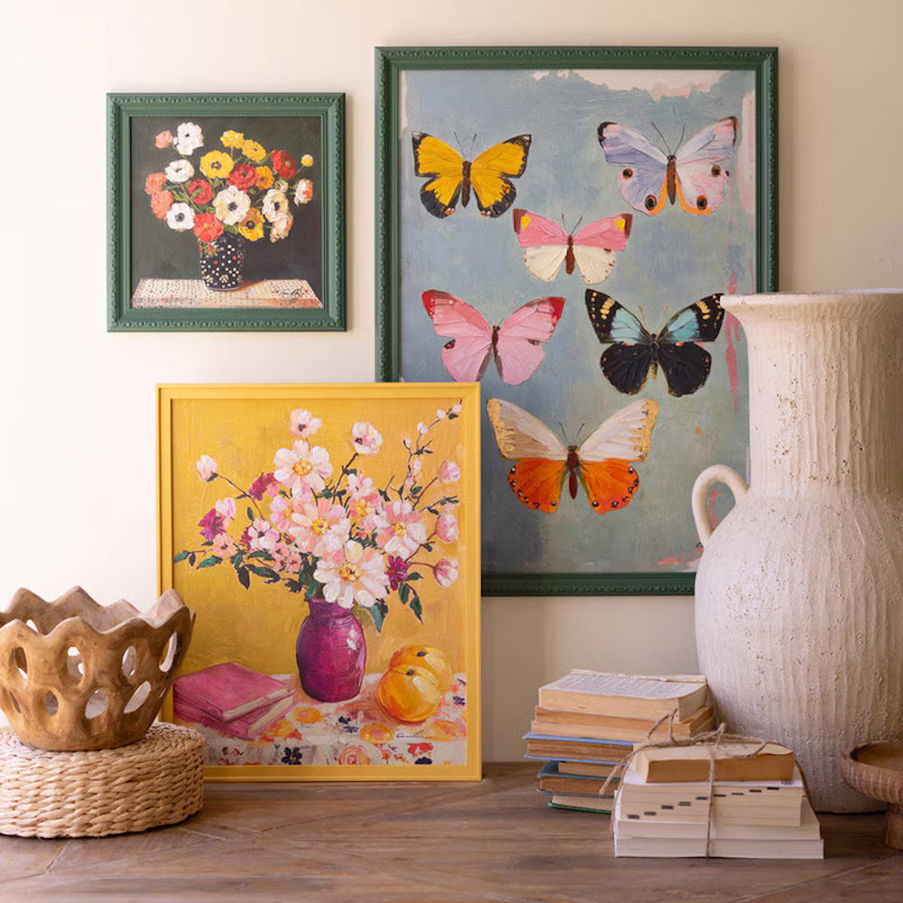 Still life art display with floral and butterfly paintings, vintage books, and a large white ceramic vase on a wood table.