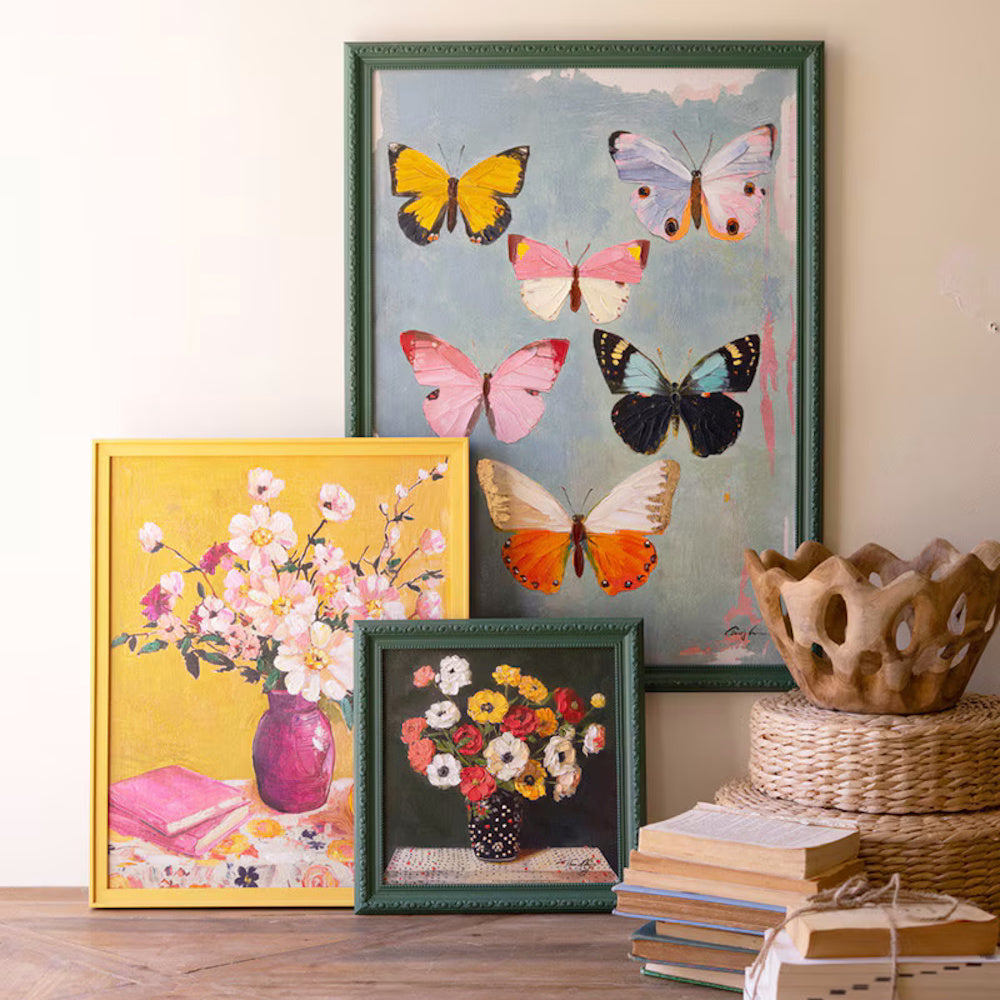 Colorful butterfly and floral still life paintings in green and yellow frames leaning against a wall with vintage books.