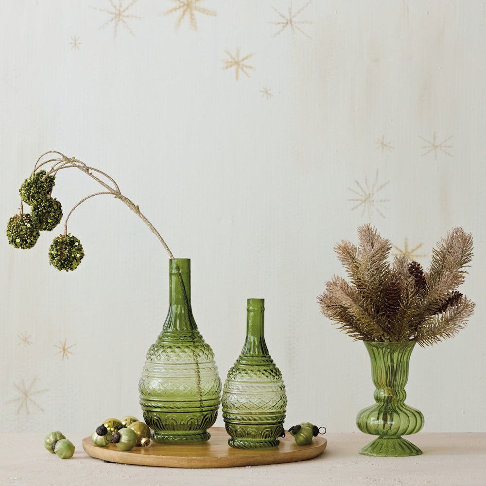 Three green glass vases display festive branches and pinecones on a table against a star-patterned wall.