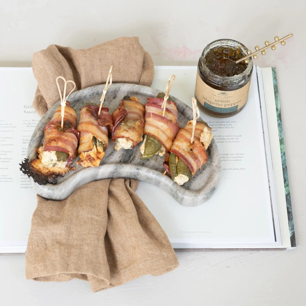 Bacon-wrapped jalapeno poppers on a stone tray next to a jar of Finch + Fennel Green Jalapeño Pepper Jelly.