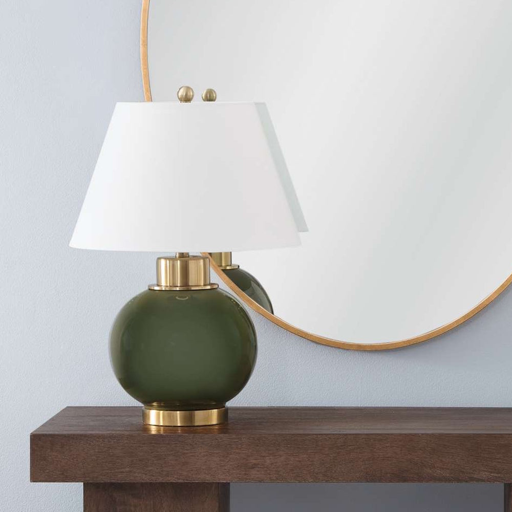 Olive green glass table lamp on a wooden table in front of a round brass mirror against a light blue wall.