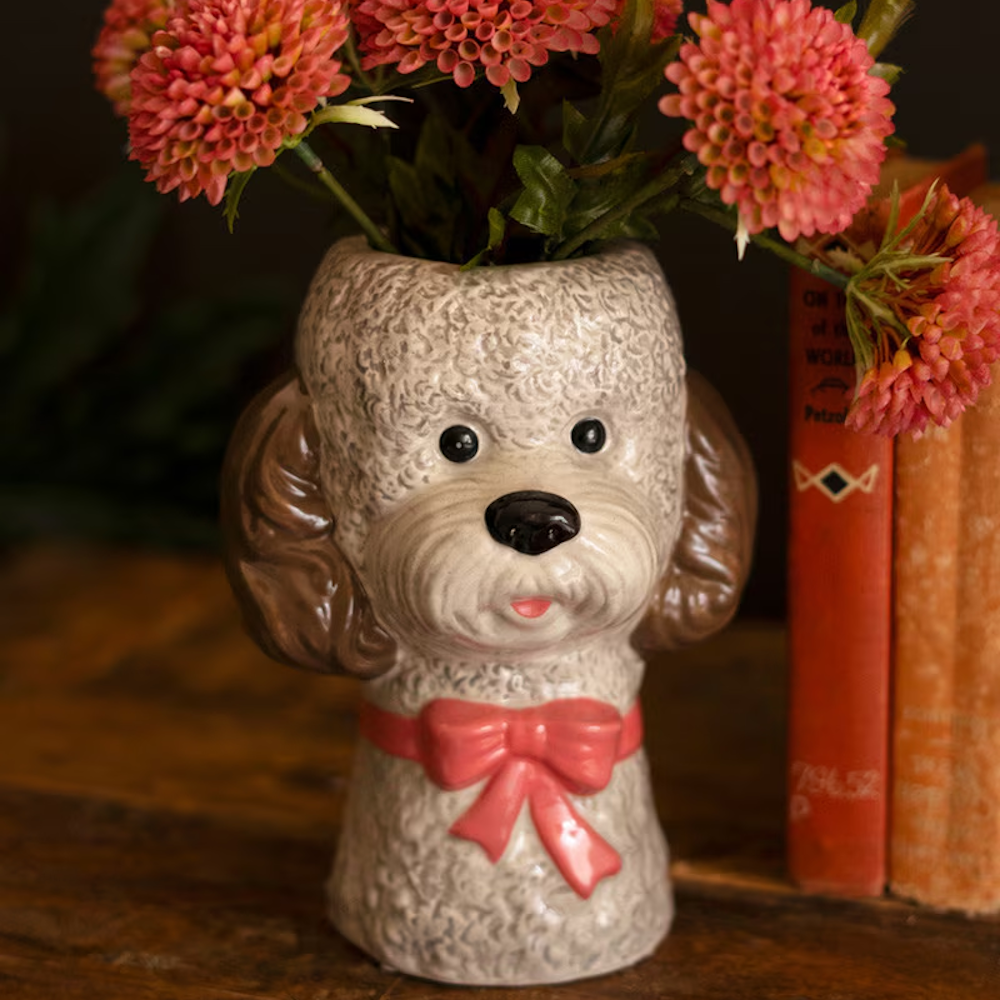 Grey ceramic poodle dog vase with pink bow and pink flowers on a wooden table.
