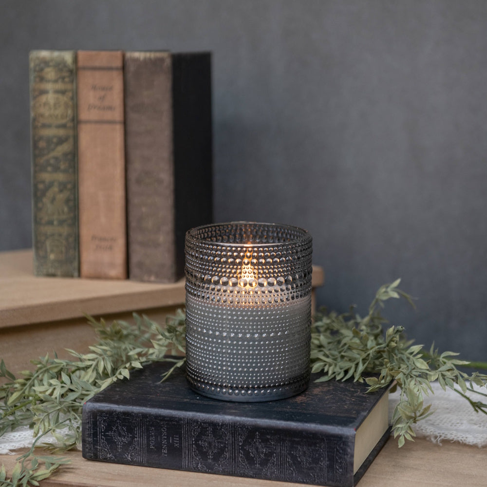 Grey dotted glass candle glowing on top of a vintage book next to greenery.