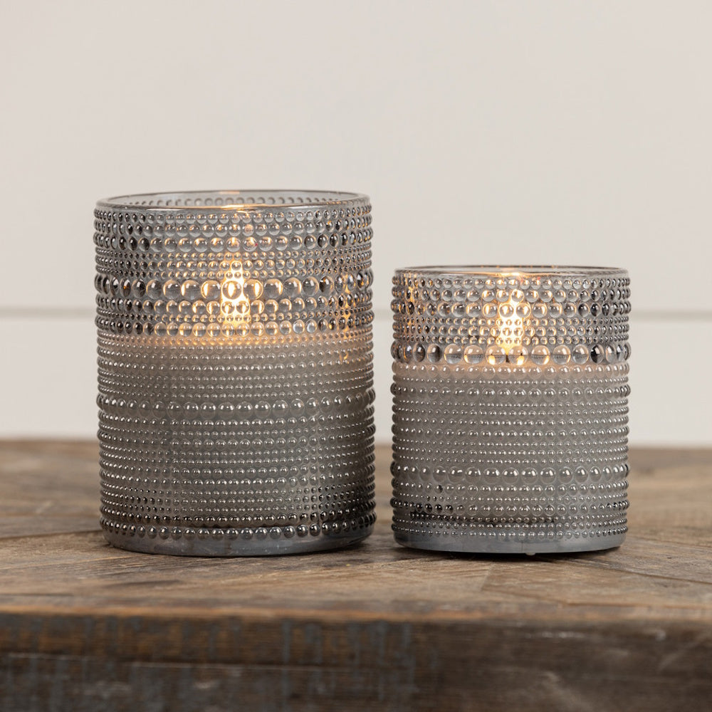 Two glowing grey hobnail glass candle holders in different sizes sitting on a wooden surface.