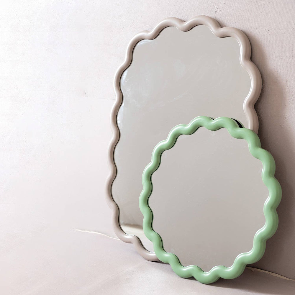 Two wall mirrors with wavy scalloped frames in neutral grey and sage green leaning against a wall.