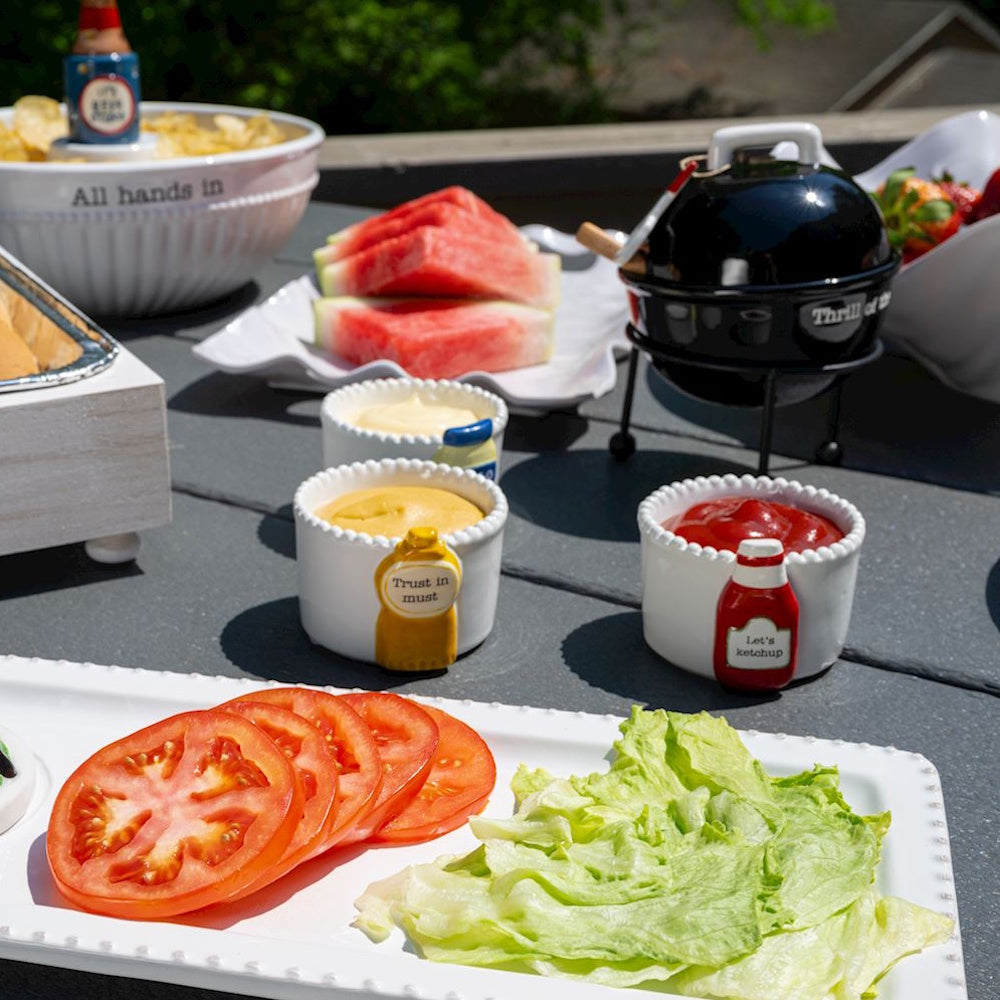 Summer cookout table with sliced tomatoes, lettuce, ketchup, mustard, and a mini BBQ grill.