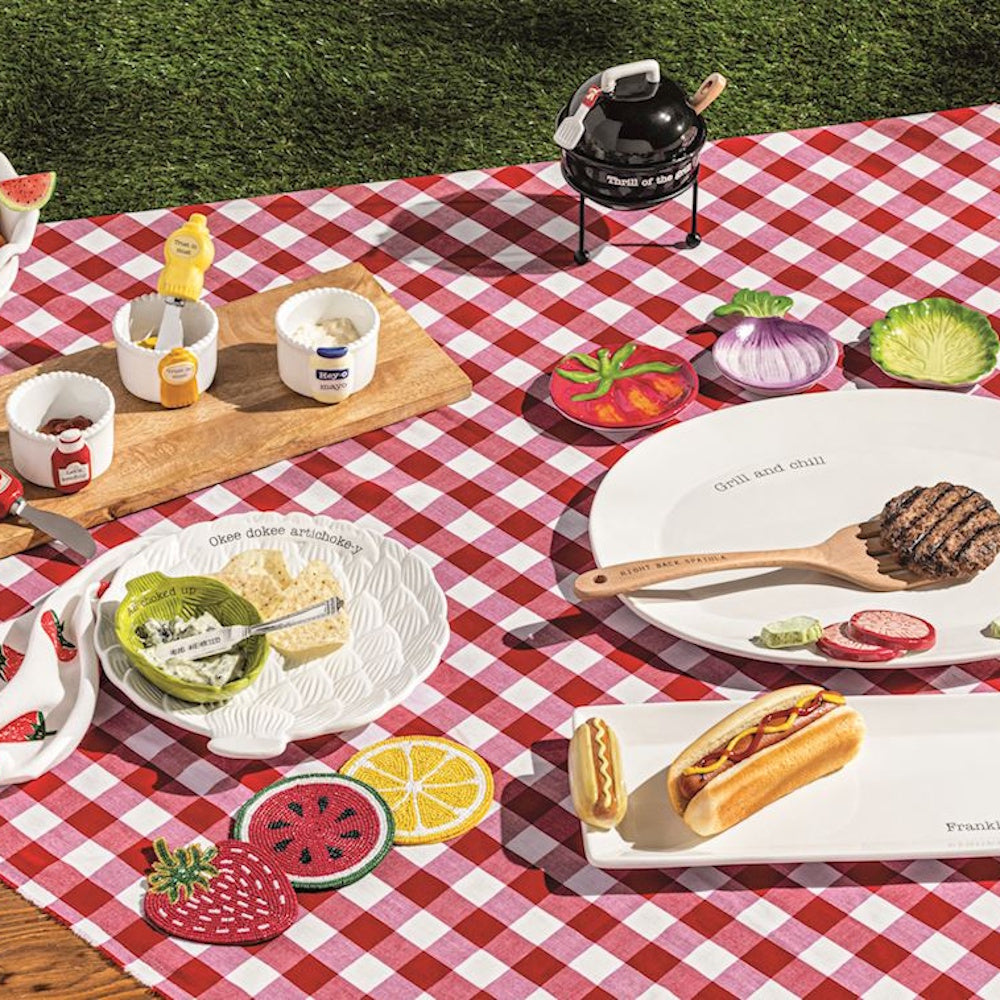 Summer picnic spread on red gingham blanket featuring grill-themed serving plates, condiment dishes, and food.