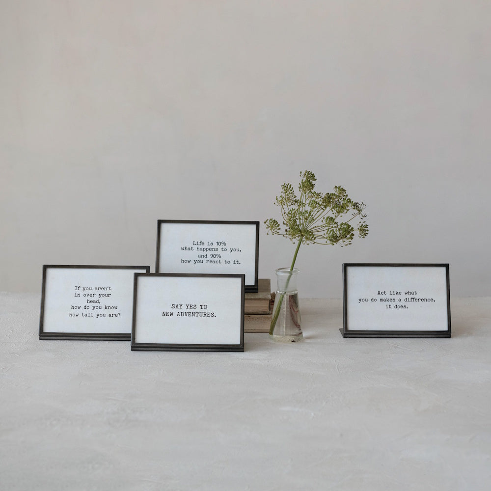 Four gunmetal finish metal and glass frames with easel backs displaying inspirational quotes, arranged on a surface with a small vase of dried botanicals.