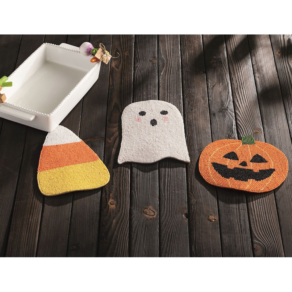 Three beaded Halloween trivets shaped like candy corn, a ghost, and a pumpkin on dark wood.