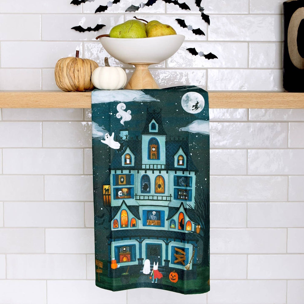Halloween kitchen towel with haunted house illustration, displayed on wooden shelf with pumpkins and bats.