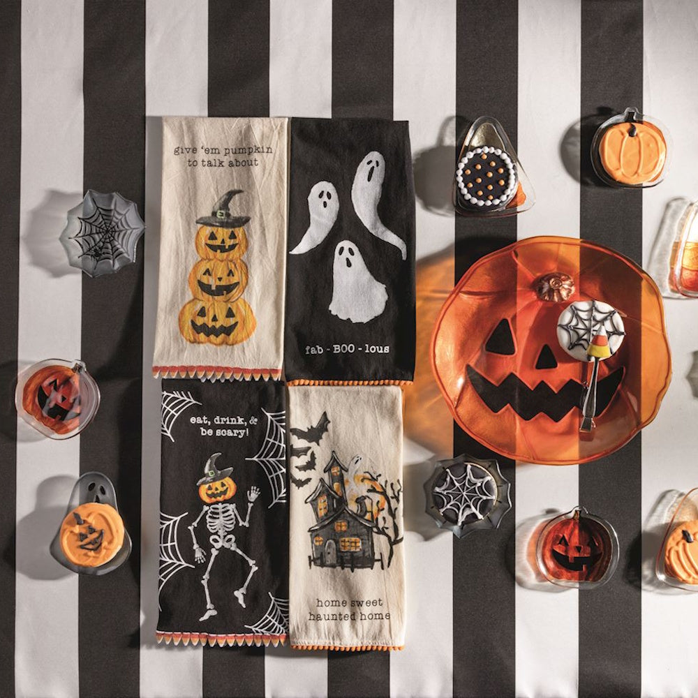 Four Halloween kitchen towels with spooky graphics on a black and white striped table with themed treats.