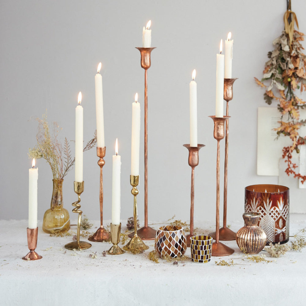 Multiple brass and copper taper candle holders with lit white candles arranged together on a tabletop display.