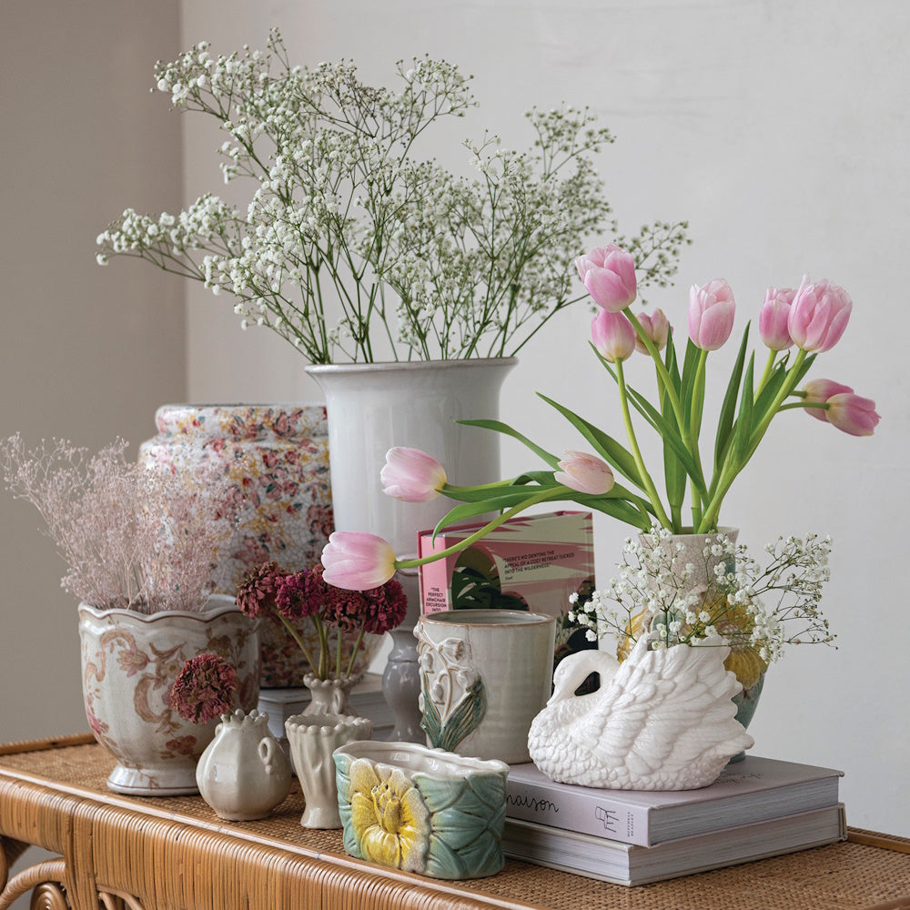 Pink tulips and baby's breath flowers arranged in a collection of decorative ceramic vases on a wicker table.