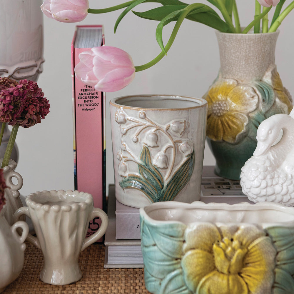 Embossed ceramic planters and vases with floral designs, books, and pink tulips on a woven surface.