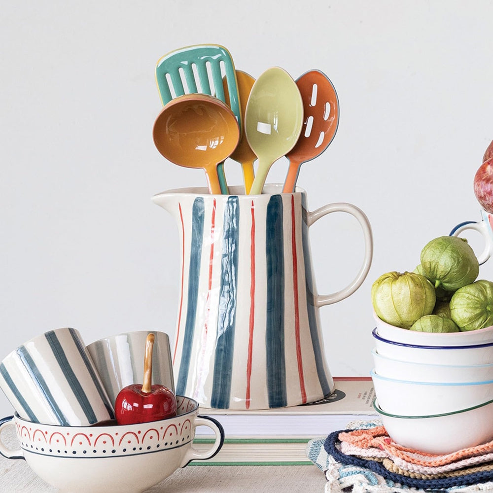 A hand-painted striped ceramic pitcher holding colorful kitchen utensils, surrounded by coordinating tableware.