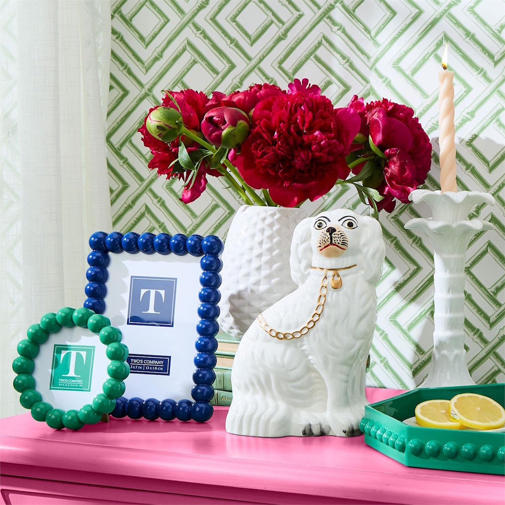 White Staffordshire dog figurine displayed on a pink table alongside frames, red flowers, and decor.