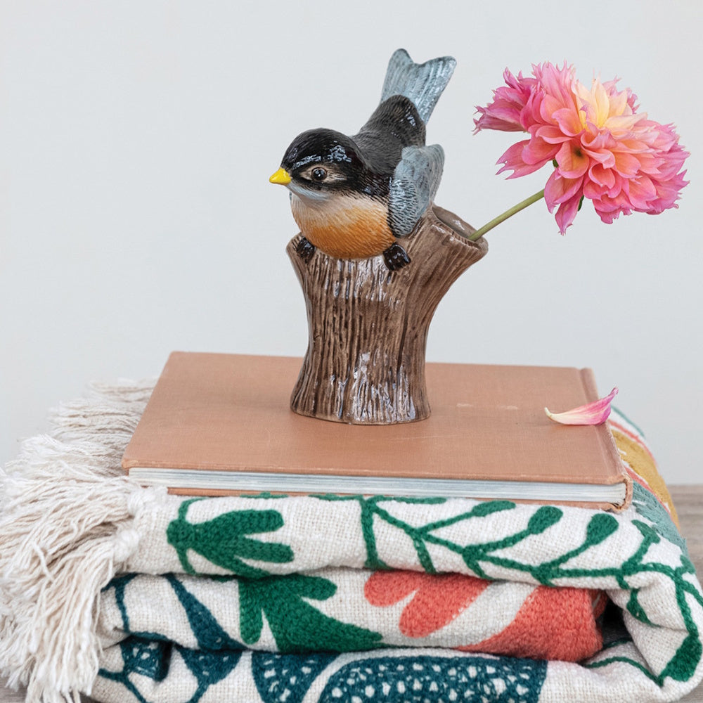 Ceramic bird figurine on a log-shaped vase holding a pink flower, sitting on a book and folded blanket.
