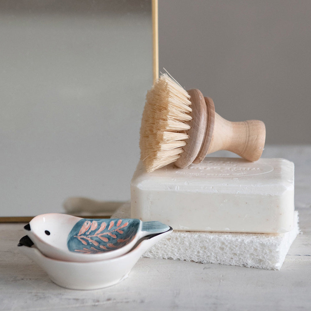 A wooden scrub brush rests on a bar of soap, next to stacked bird-shaped ceramic trinket dishes.
