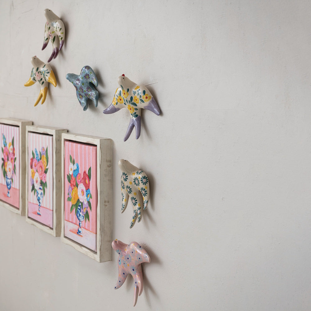 Hand-painted ceramic birds and framed floral paintings mounted on a textured, off-white wall.