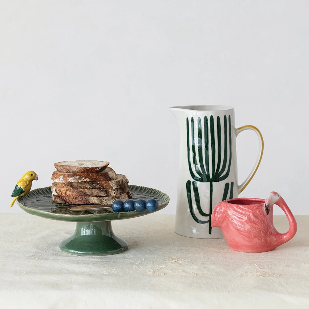 Hand-painted stoneware flamingo mug, green ceramic cake stand with bread, and floral pitcher on a neutral tabletop.