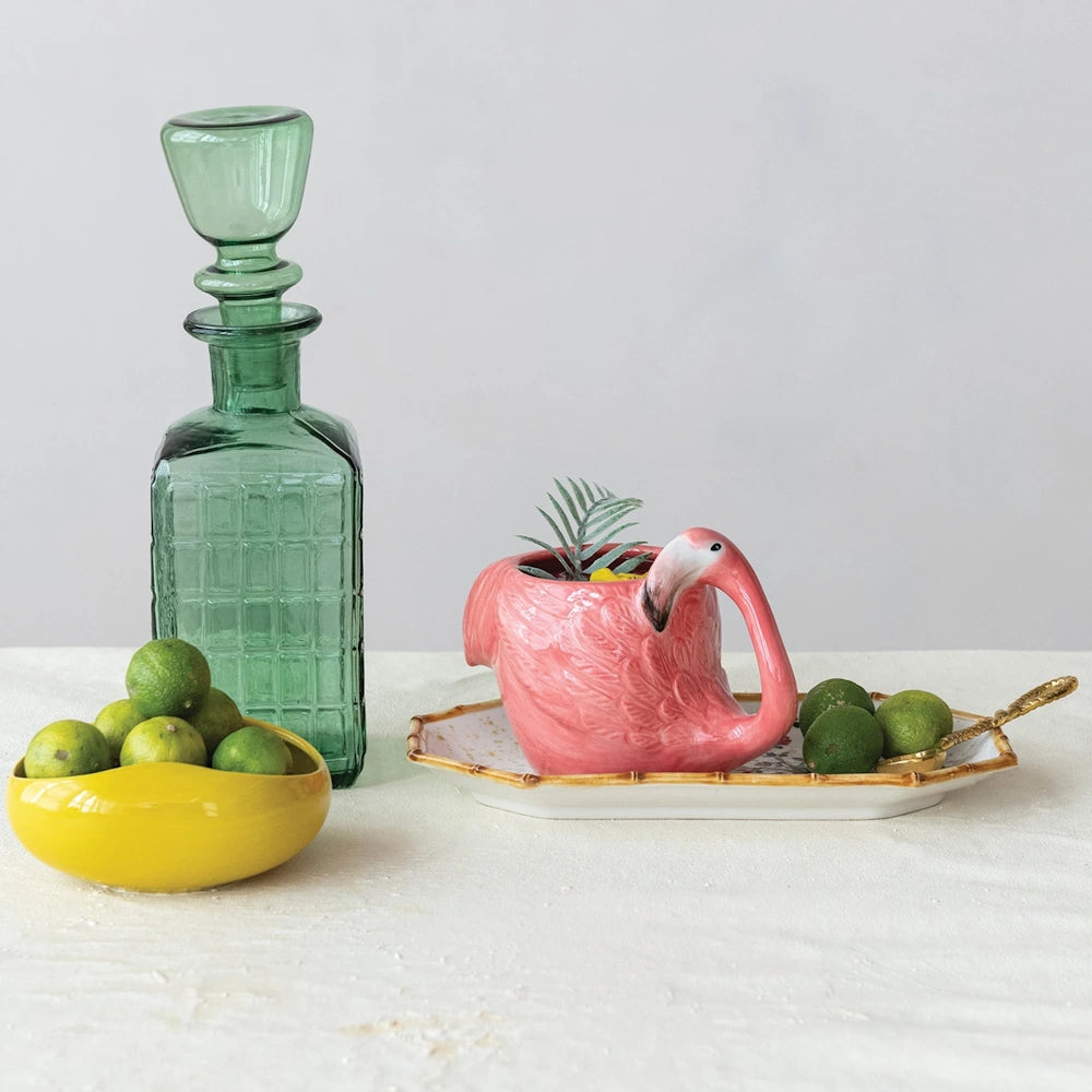 Hand-painted pink flamingo stoneware mug on a tray with green limes, a yellow bowl, and a vintage green glass decanter.