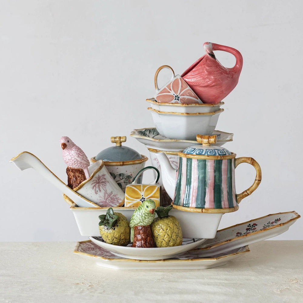 Stacked hand-painted tropical stoneware featuring a flamingo mug, bird figurines, and citrus-patterned teapots and bowls.