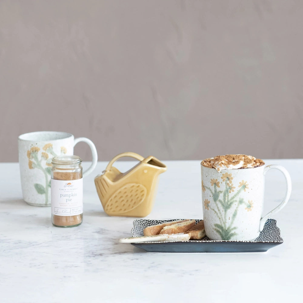 Botanical stoneware mug filled with a whipped hot drink, served alongside biscotti, spice, and a creamer.