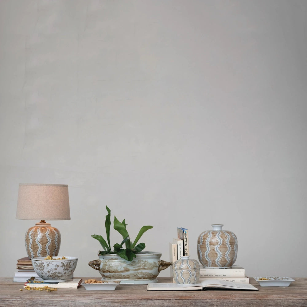 Lifestyle view of the lit ceramic table lamp on a wooden console table alongside matching patterned pottery and books.
