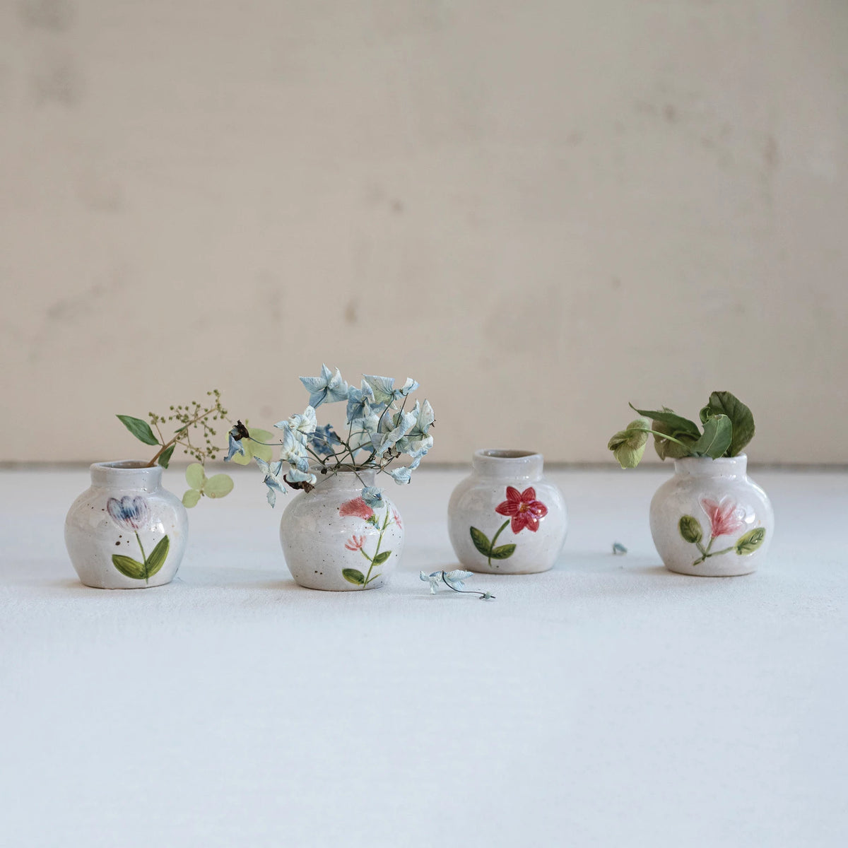 Four small hand-painted stoneware bud vases with floral designs and dried flowers on a neutral white surface.