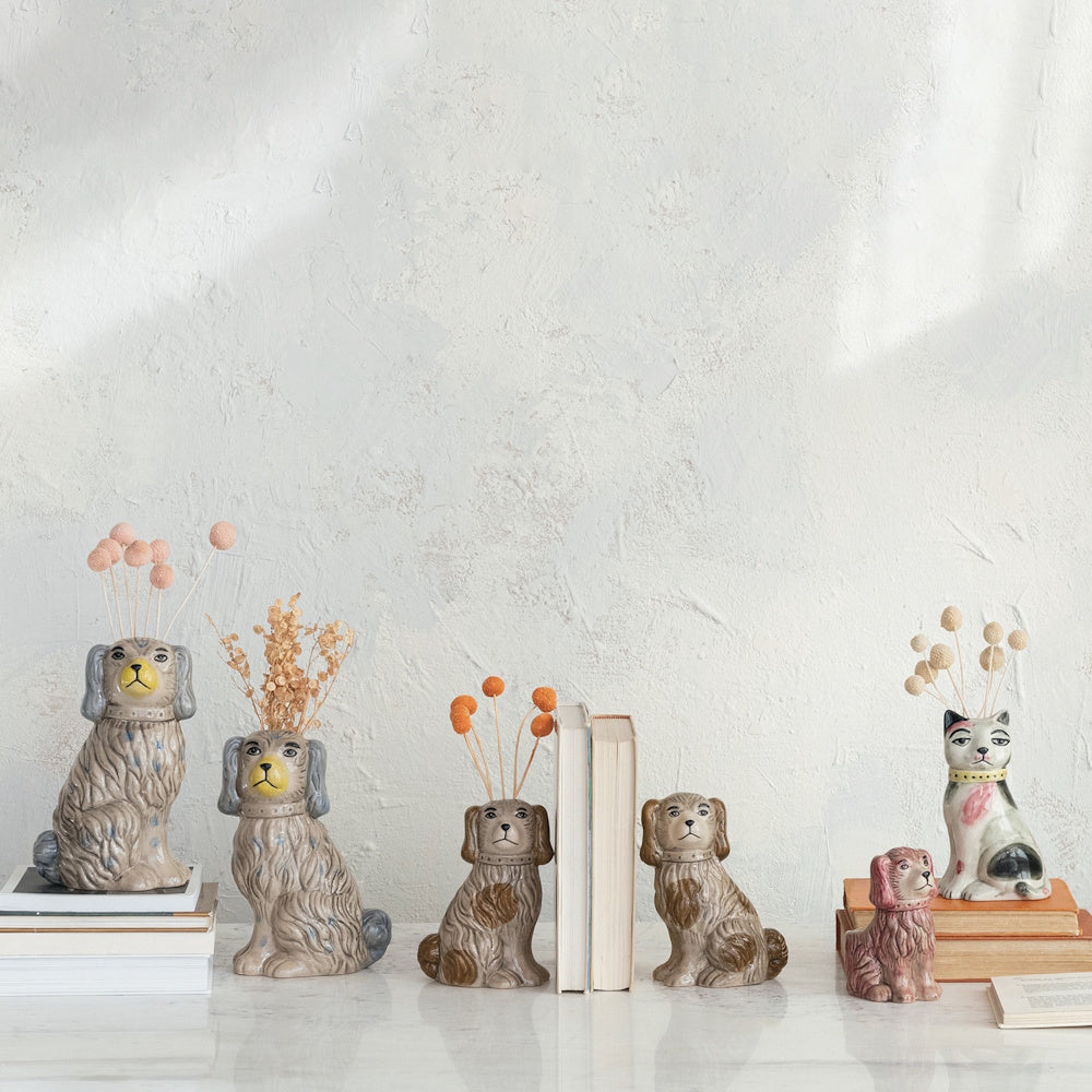 Collection of hand-painted vintage-style dog and cat ceramic vases with dried flowers arranged on books and a white ledge.
