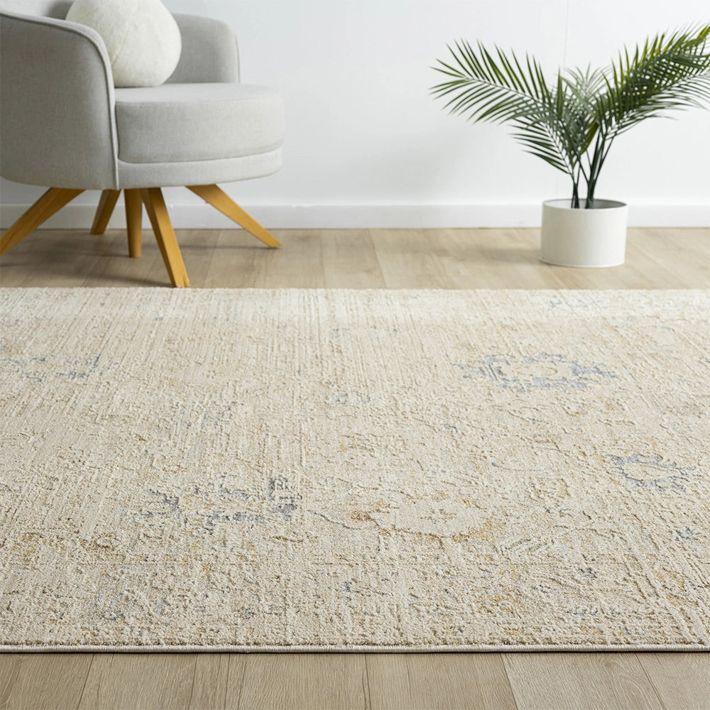 Beige Monaco rug with a distressed blue and gold pattern in a modern living room with a grey chair and palm plant.