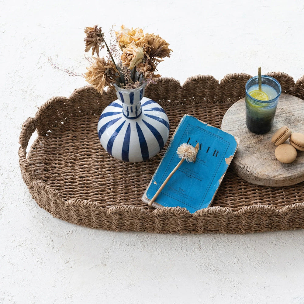 Woven scalloped tray with a vase of dried flowers, a blue book, and a drink with macarons.