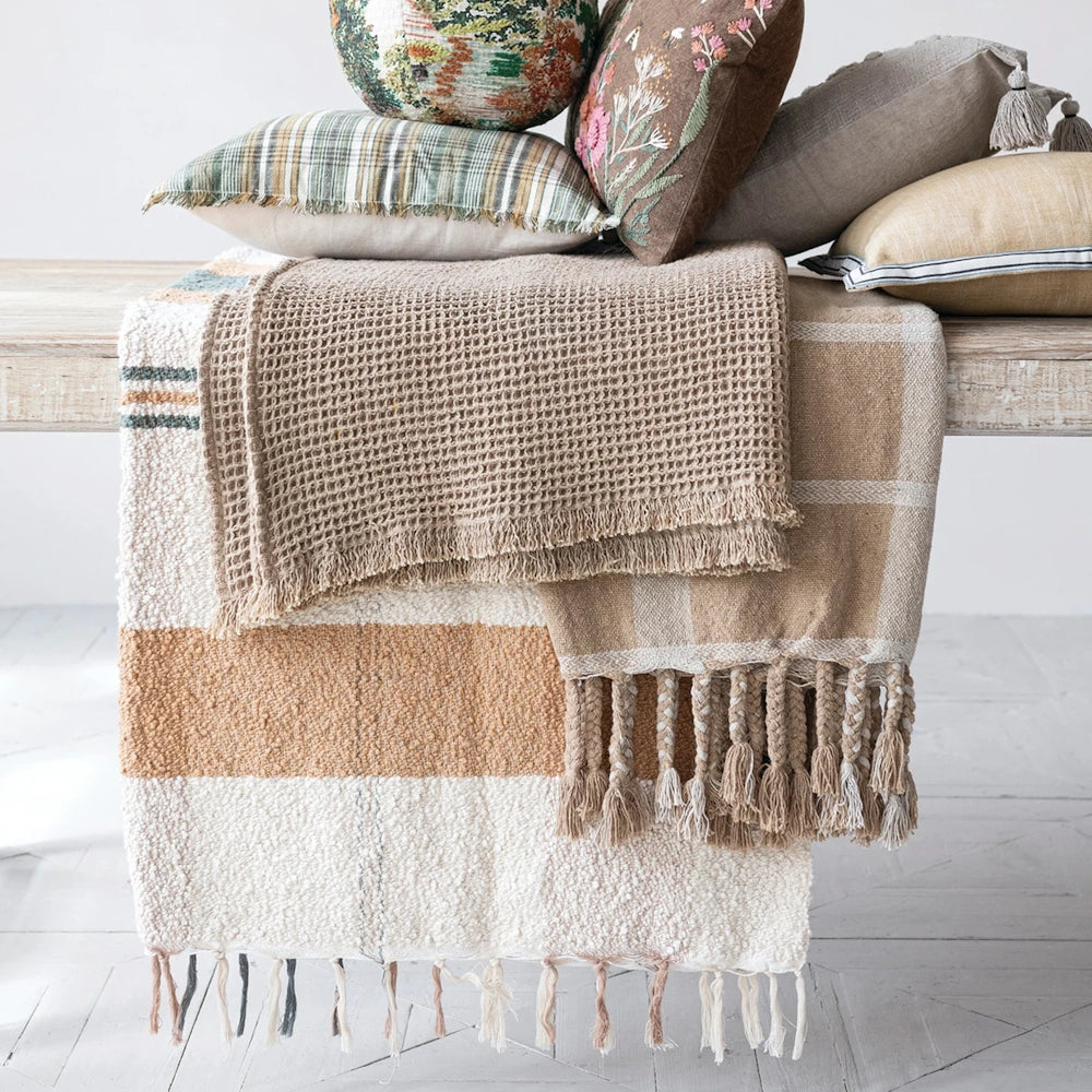 Lifestyle view of multi-colored woven cotton throw blankets with fringe, styled on a bench under throw pillows.