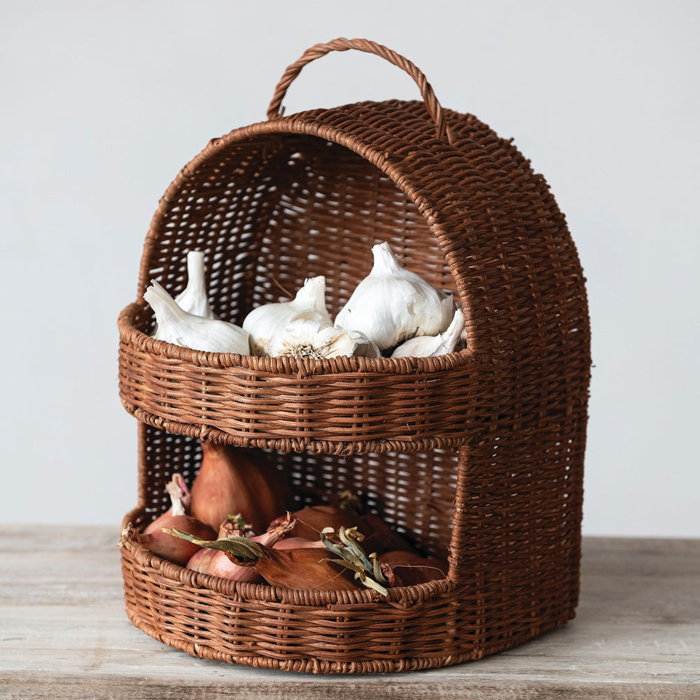 Hand-woven rattan 2-tier basket with handle holding garlic and shallots on a wooden table.