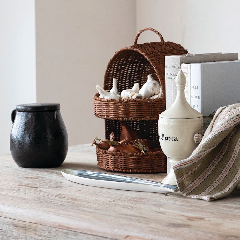 Hand-woven rattan 2-tier basket with handle holding garlic and shallots on a rustic wooden table with kitchen decor.