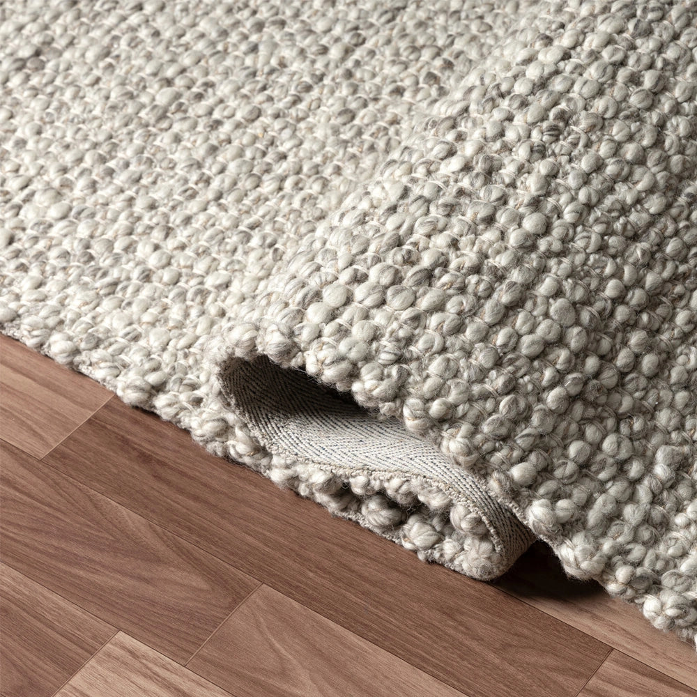 Hand-woven rectangle dome rug with thick textured grey wool loops rolled up on a wood floor.
