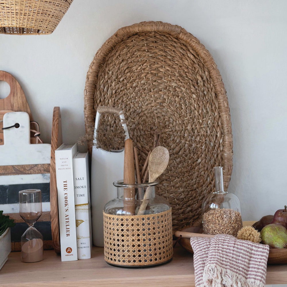 Oval woven seagrass tray displayed on a kitchen counter with cookbooks, wooden utensils, and glass jars.