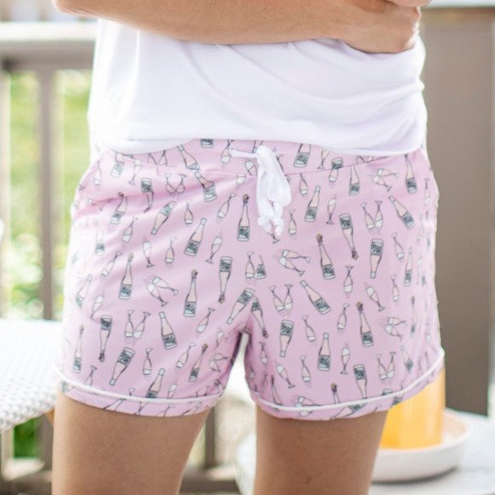 A person wearing pink pajama shorts with a champagne bottle and glass pattern and a white t-shirt.