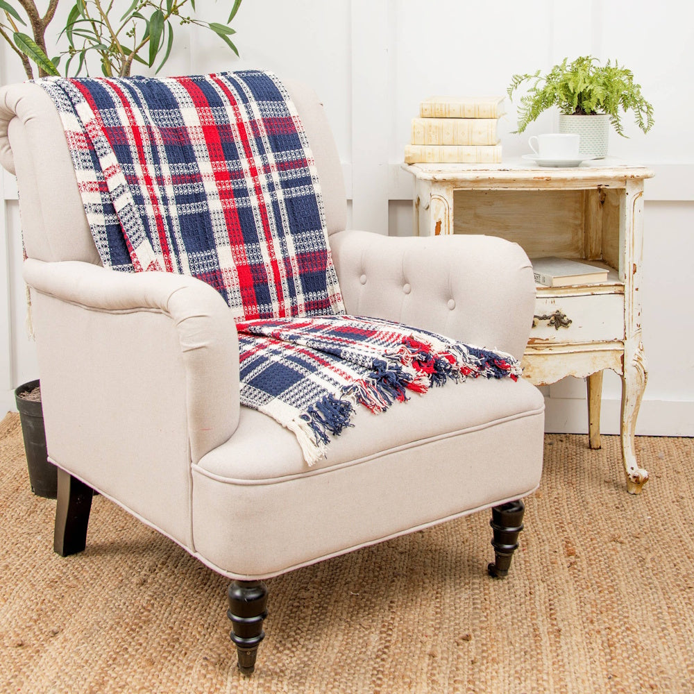 Red, white, and blue plaid throw blanket with fringe draped over a tufted beige armchair next to a side table.