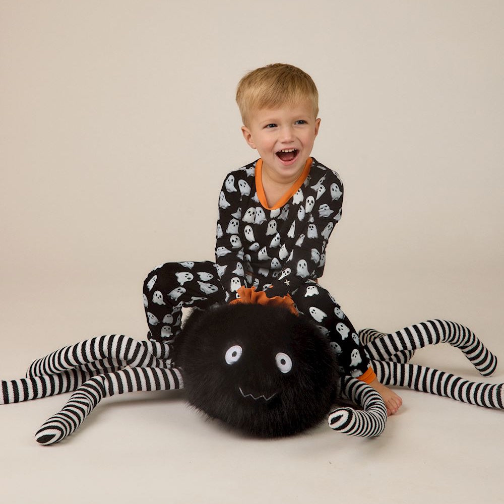 A toddler in ghost pajamas sitting on a large, plush black spider with striped legs.