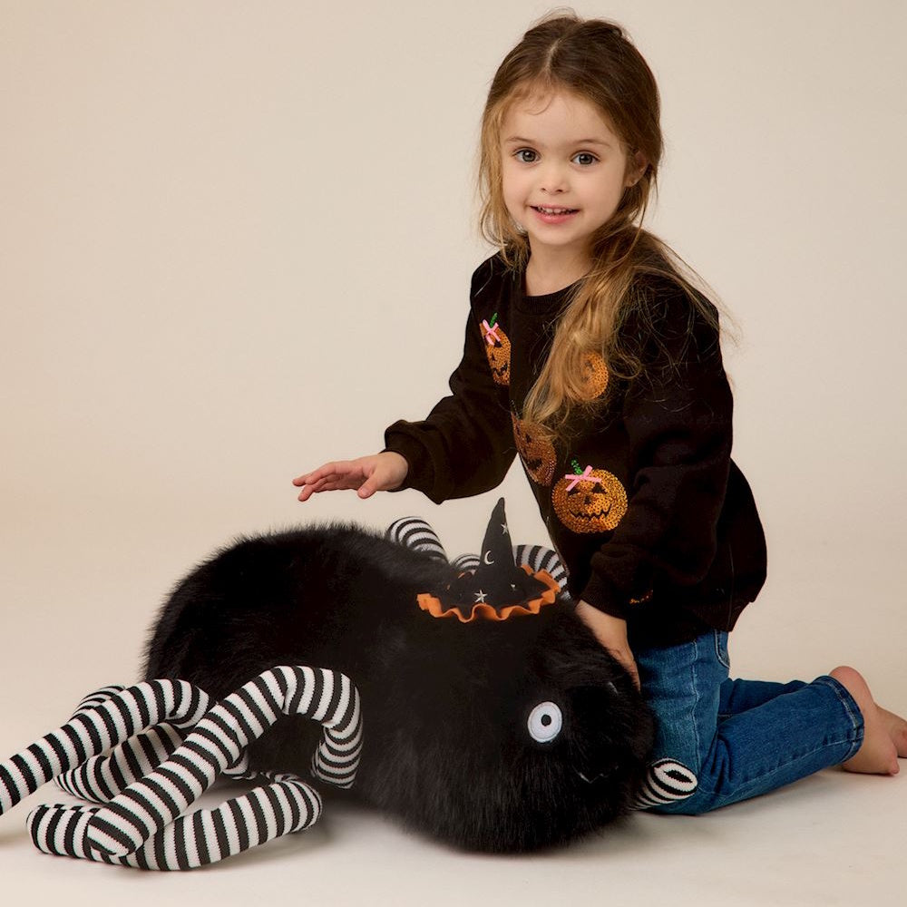 Young girl playing with a large plush black spider featuring striped legs and a mini witch hat.