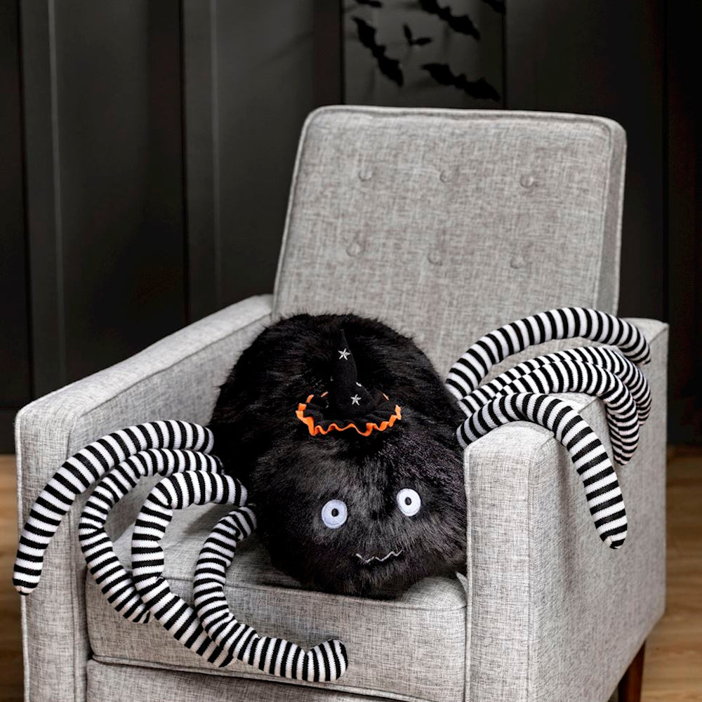 Plush black spider toy with striped legs wearing a witch hat, sitting in a grey armchair.
