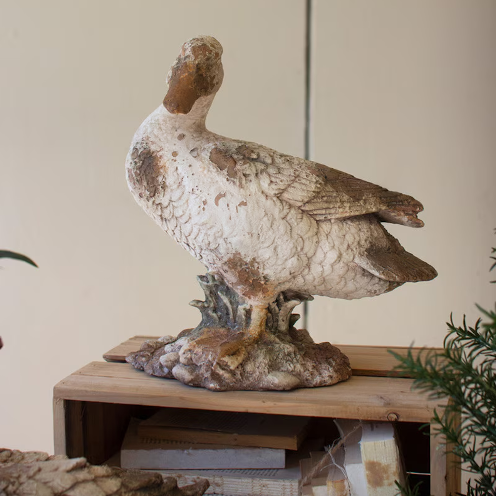 Weathered duck statue with head turned, sitting atop a wooden crate filled with books.