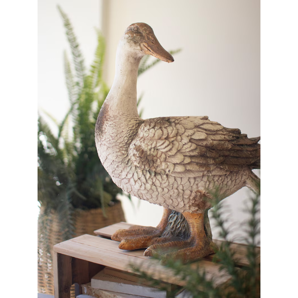 Rustic standing duck statue with neck upright, displayed on a wooden crate next to potted plants.