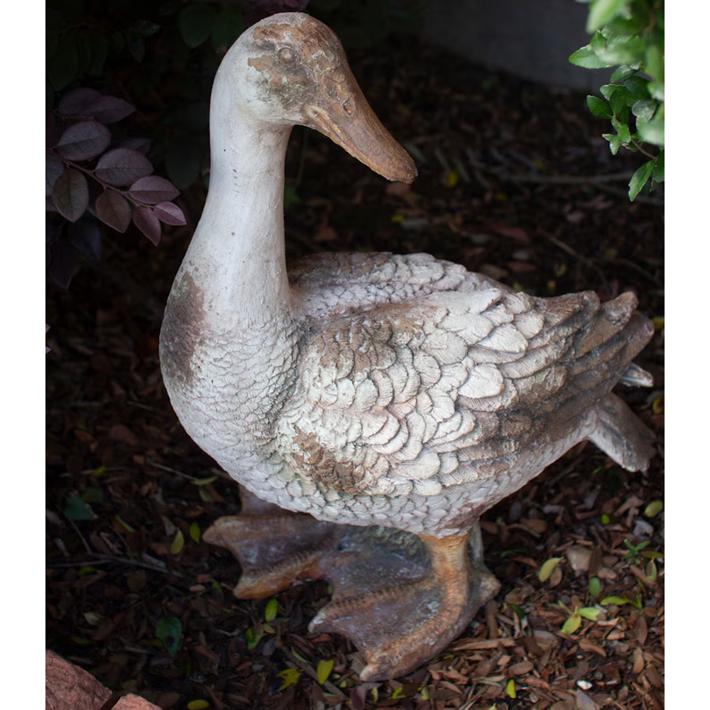 Faux-concrete duck garden statue standing with head held up amidst dark soil and foliage.