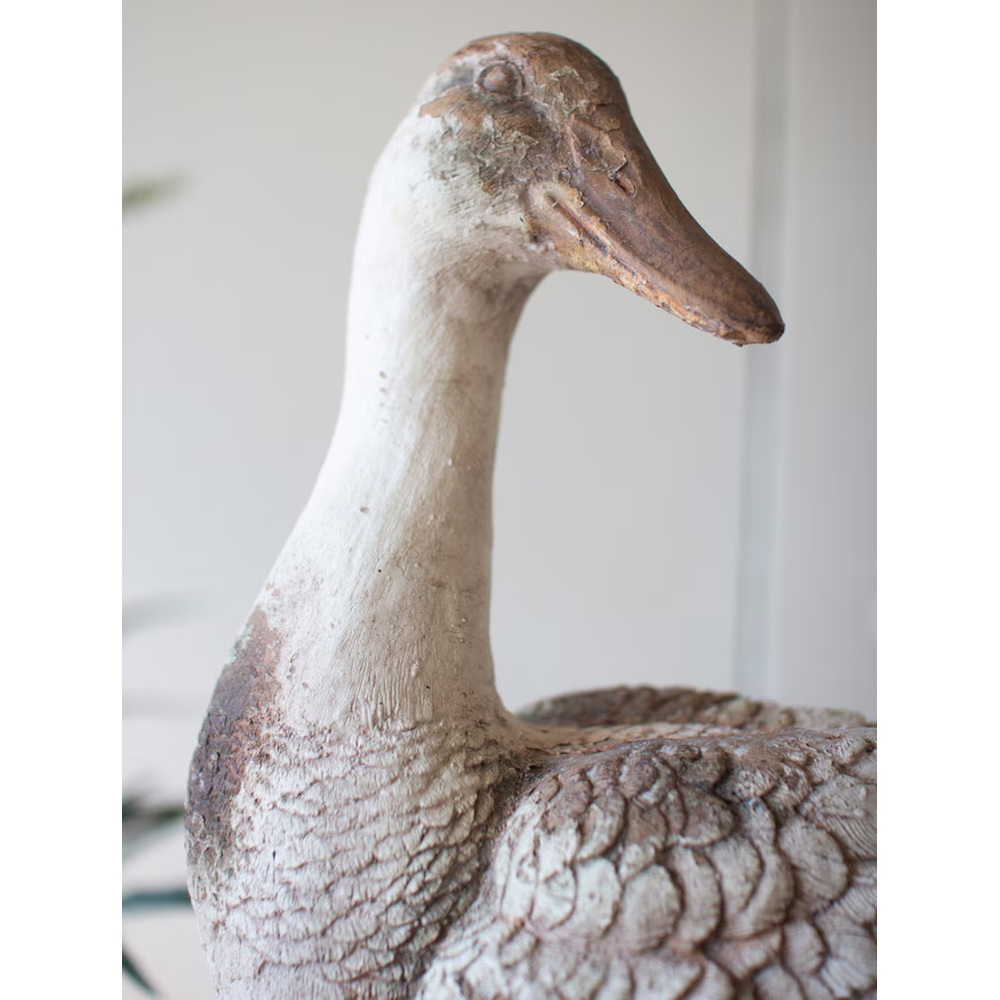 Close-up of a faux-concrete duck statue with a weathered white and brown finish against a plain wall.