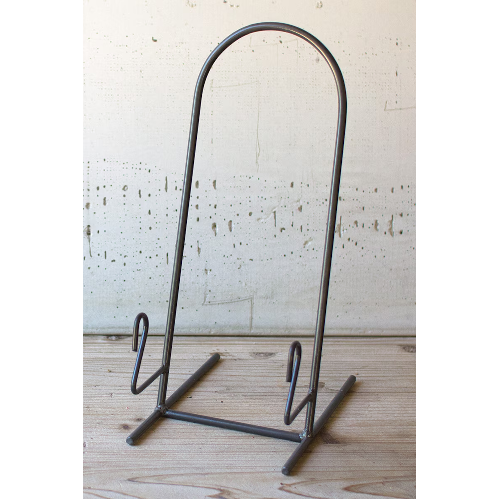 Minimalist dark iron plate easel display stand resting on a light wooden tabletop.