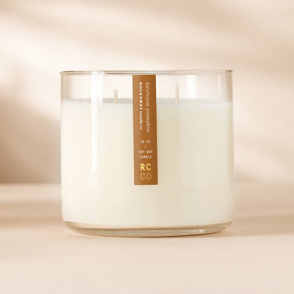 A white soy wax candle in a clear glass jar with a brown label, set against a neutral, warm background.